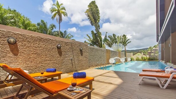 A poolside area with lounge chairs, a table with a glass of wine, and a stone wall. - Chic Condo Karon by RESAVA (Karon)
