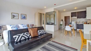 A modern living room with a sofa, a dining area with a table and chairs, and a kitchen in the background. - Chic Condo Karon by RESAVA (Karon)