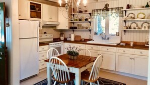A kitchen with white cabinetry, a wooden dining table with two chairs, a chandelier, and a window with a plaid curtain. - Sea Rose Cottage - Cozy, Comfortable, Ocean Views on Orr's Island, Harpswell, ME (Harpswell)