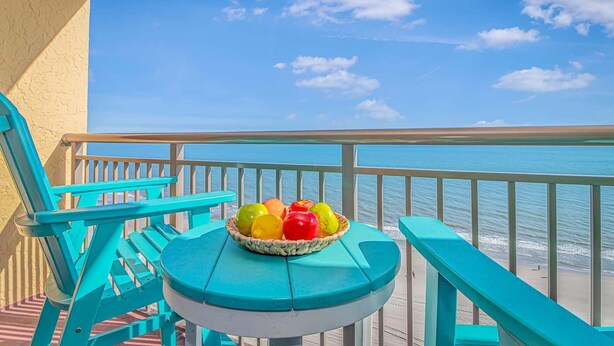 A turquoise deck with a table holding a basket of fruit, overlooking a beach with clear blue water and a few clouds in the sky.