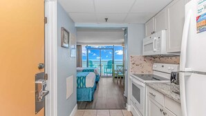 A narrow hallway leading to a balcony with a view, a kitchenette with a microwave, and a bed with blue bedding.
