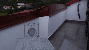 A balcony with a metal table and chair, a red railing, and a view of a hilly town.
