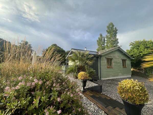 A small, single-story house with a sloped roof, surrounded by a gravel driveway and various plants. - Kingfisher Lodge - Private Hot Tub & Parking (Exeter)