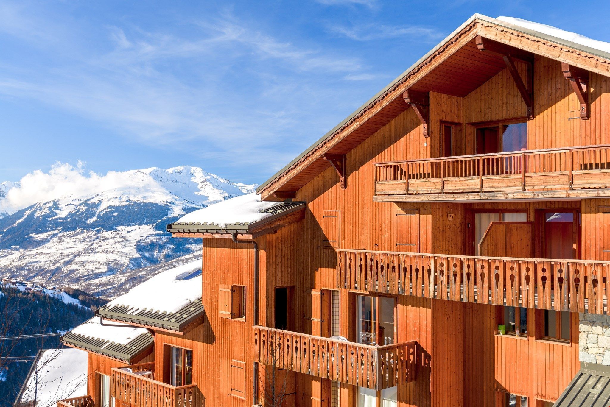 A wooden chalet with balconies, a snow-covered mountain backdrop, and a clear sky.