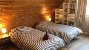 A cozy bedroom with two beds, wooden sloped ceiling, and a small shelf with decorative items.