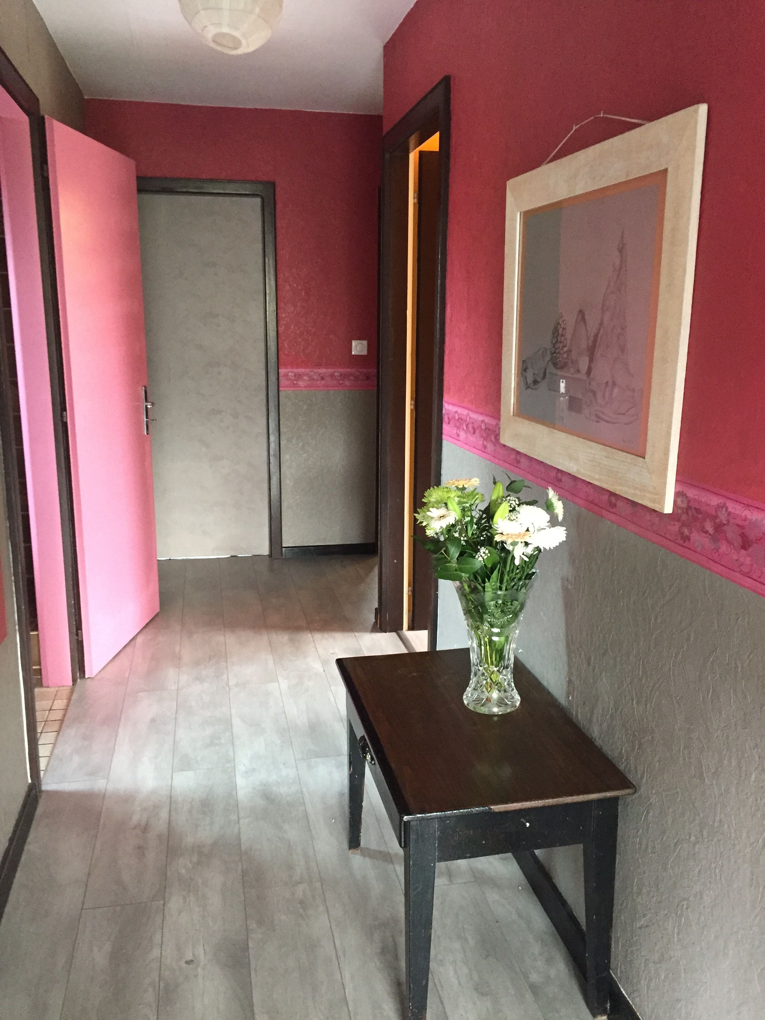 Een hal met een houten tafel, een bloemenvaas, een deur en een lijst met een afbeelding aan de muur.