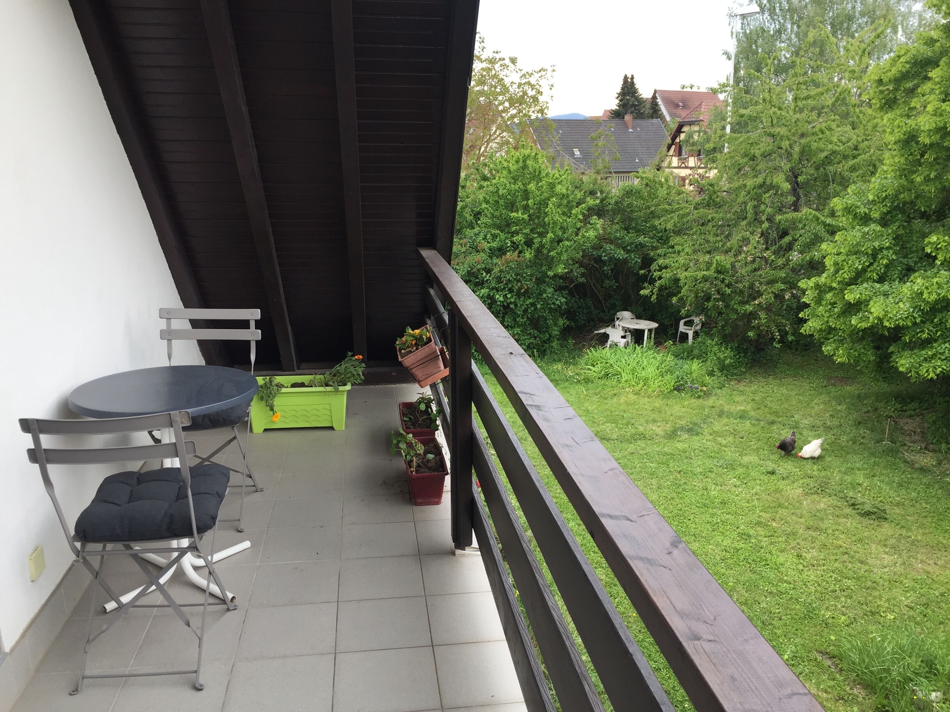 A balcony with a table and chairs, potted plants, and a view of a grassy area with trees.