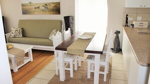 Ein Essbereich mit einem Holztisch, weißen Stühlen und einer gewebten Tischmatte. Im Hintergrund befindet sich ein grünes Sofa und ein kleiner Tisch mit einer gewebten Abdeckung.