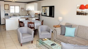 A modern kitchen with white cabinets, a dining area with a table and chairs, and a living room with a sofa and armchairs. - Santini Village 103 (Plettenberg Bay)