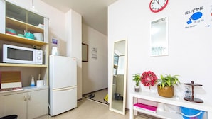 Een kleine keuken met een magnetron, koelkast en kastjes. Er is een spiegel, een klok en een bord aan de muur.