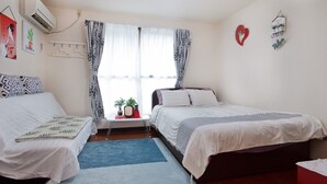 Een slaapkamer met twee bedden, een tafeltje met een plant en een radio, een raam met gordijnen met een patroon, en een wandairconditioner.