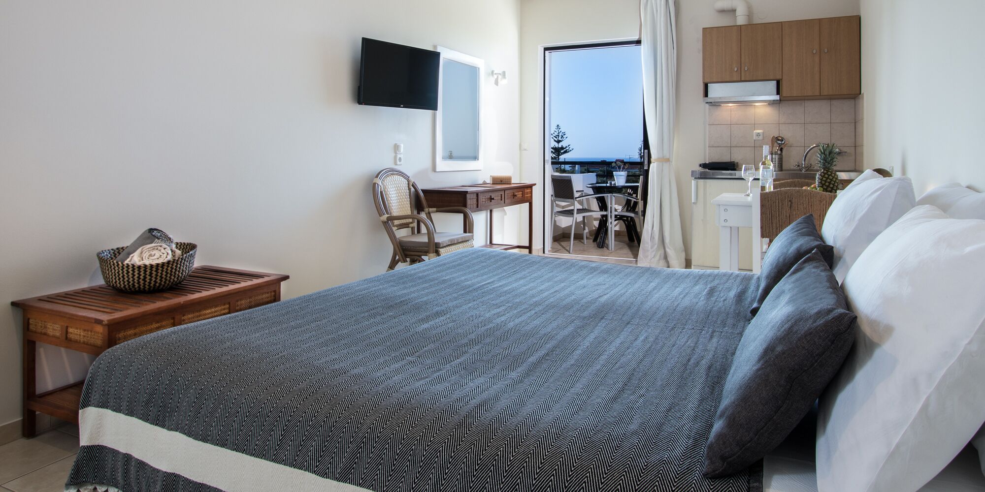 Een slaapkamer met een groot bed, een houten nachtkastje, een stoel, een bureau en uitzicht op de keuken en het balkon.