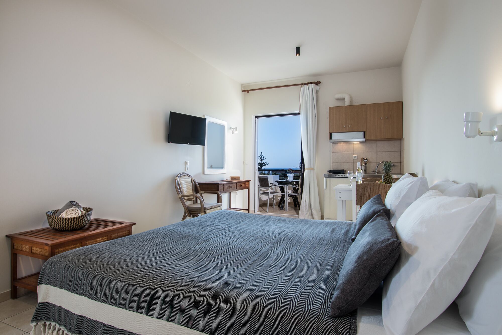 Ein Schlafzimmer mit Bett, Schreibtisch, Stuhl, Fernseher und Blick auf Balkon und Küche.