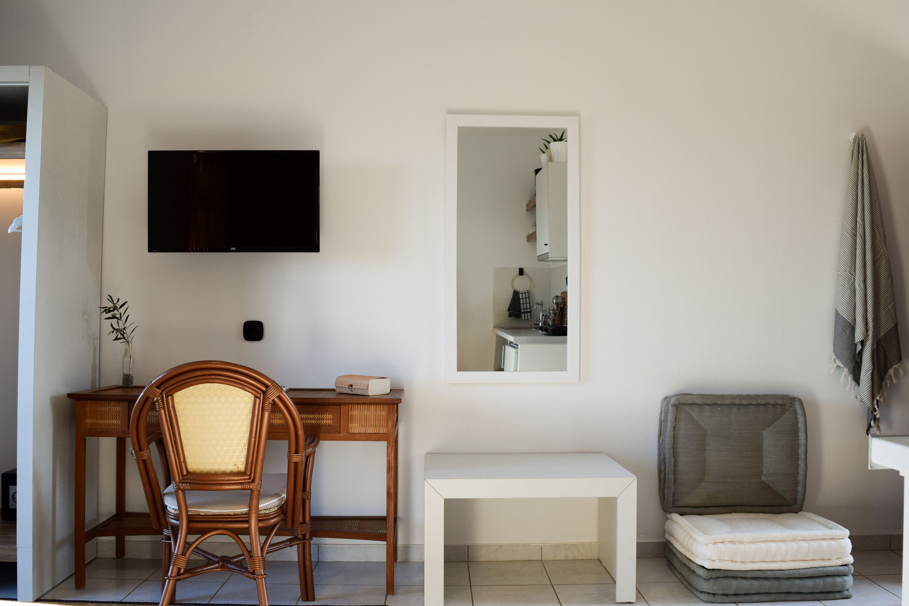 Une pièce comprenant un bureau en bois, une chaise en osier, un banc blanc, une chaise rembourrée grise, un téléviseur fixé au mur et un miroir qui reflète une salle de bain.
