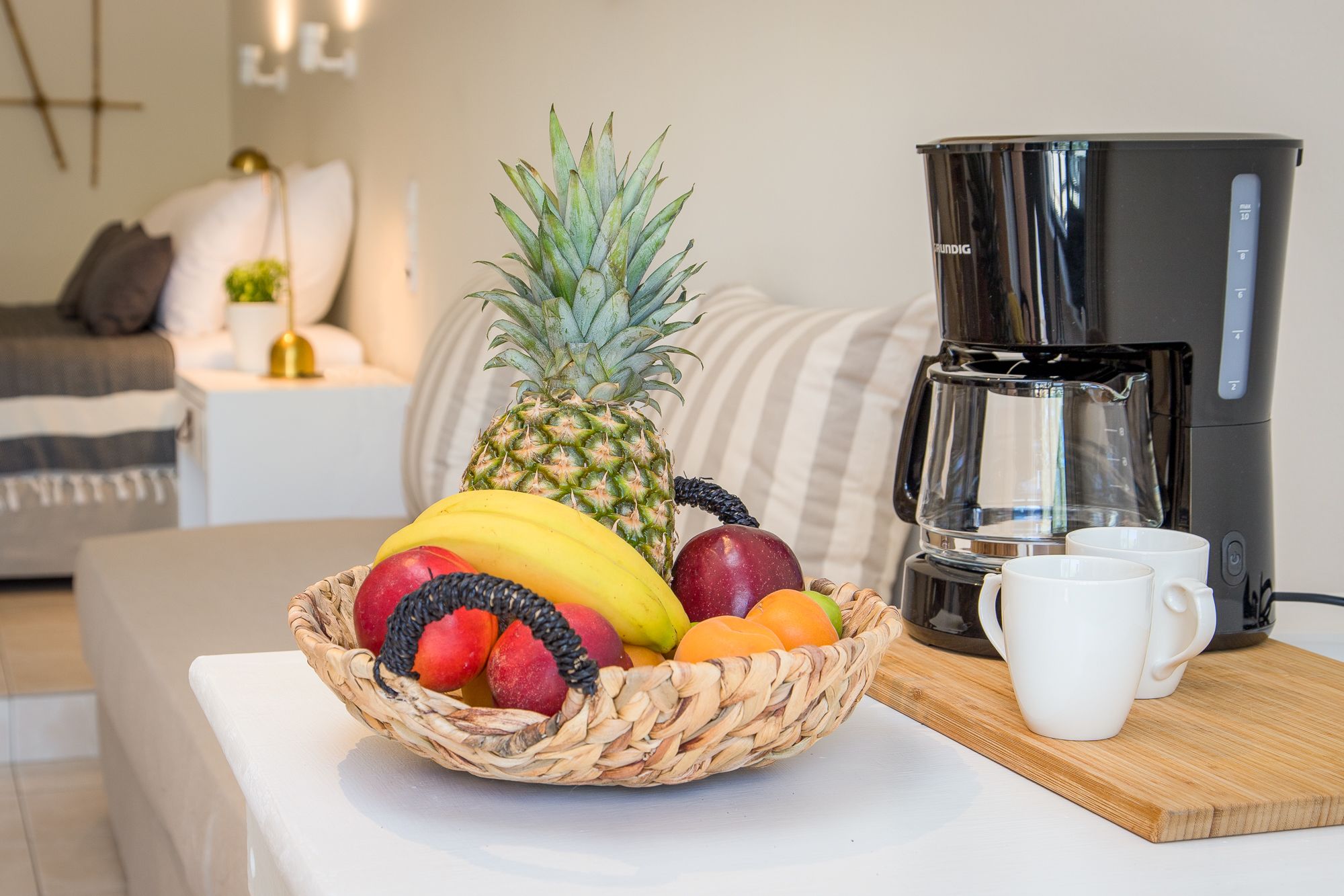 Una cesta de frutas sobre una mesa con una cafetera y dos tazas.
