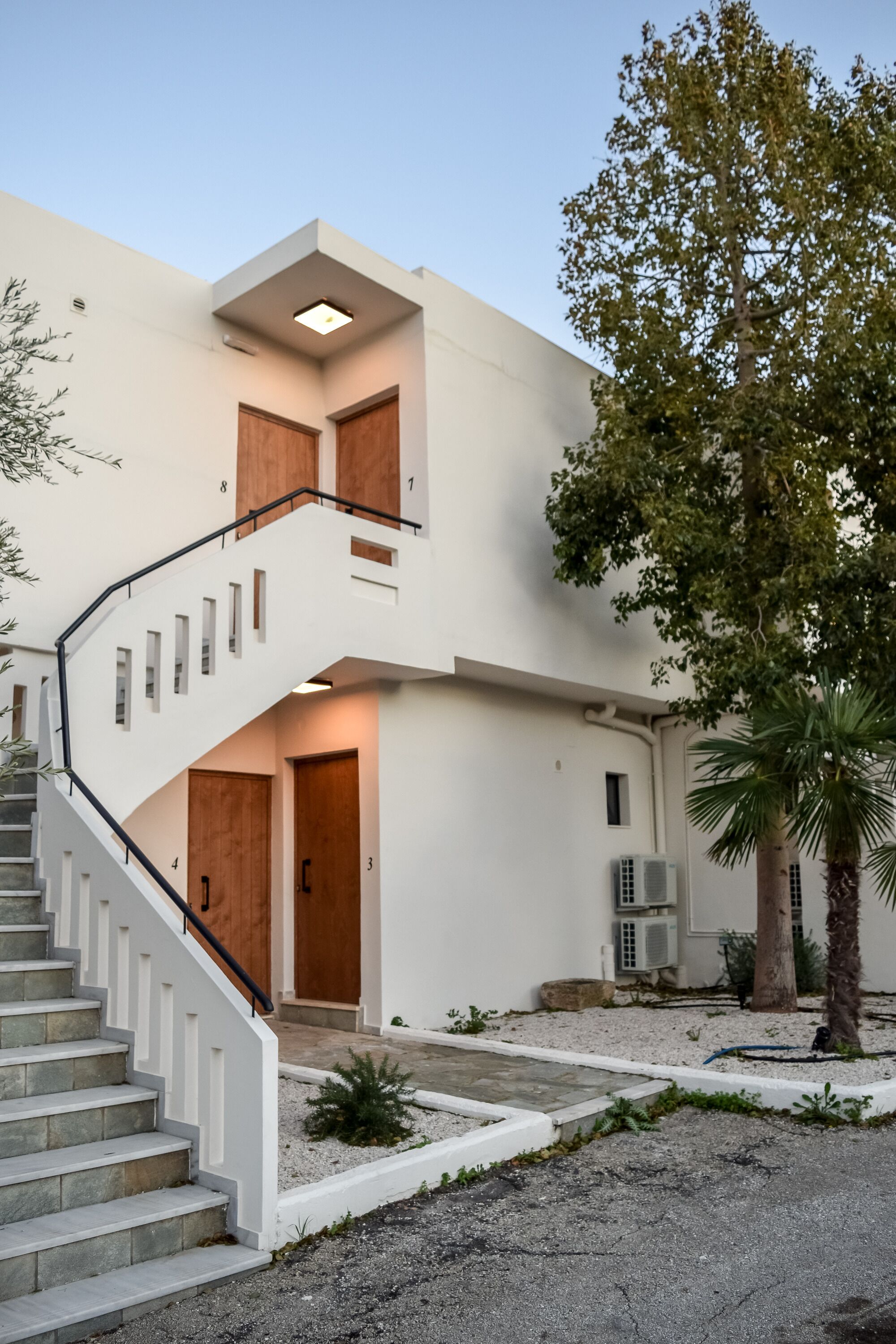 A modern two-story building with a white exterior, a staircase leading to the upper floor, and a small garden area with a palm tree.