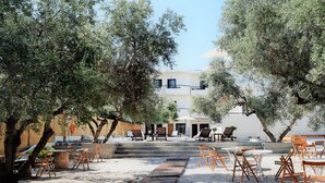 An outdoor seating area with wooden tables and chairs, surrounded by olive trees, leading to a building with multiple balconies. - Kanso Rooms (Chania)