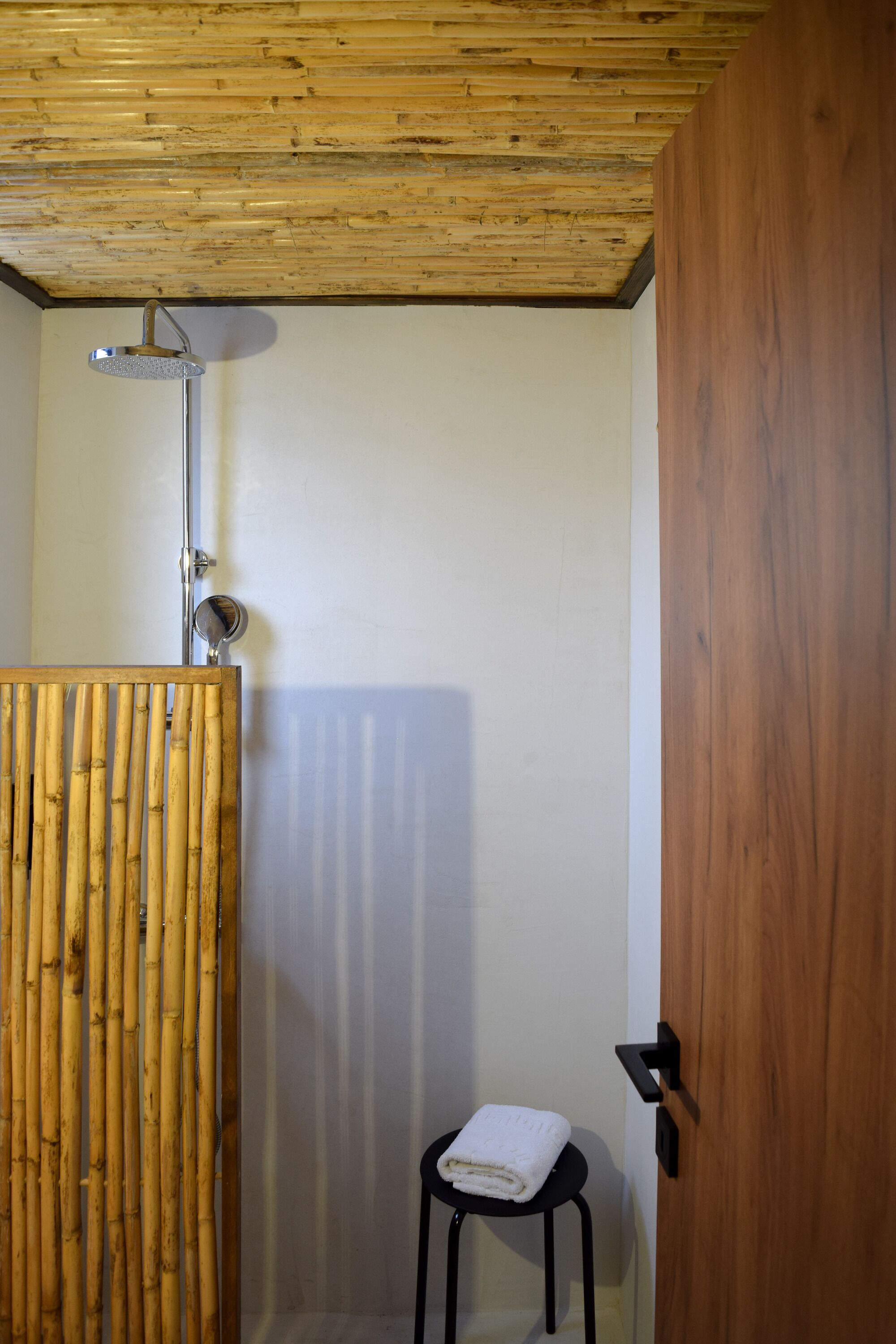 A bathroom with a wooden ceiling, a bamboo partition, a shower, and a small stool with a towel.