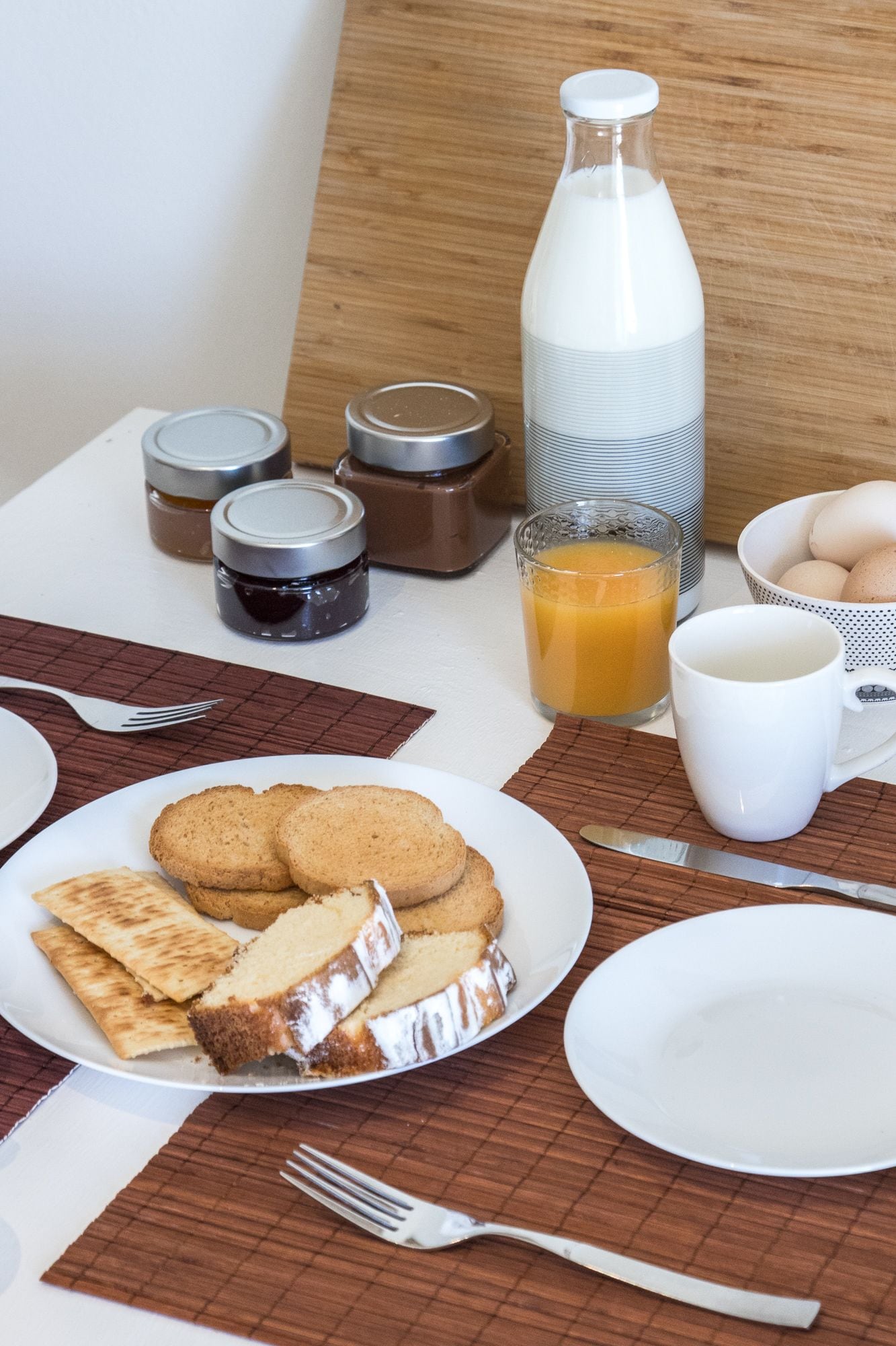 Une table dressée pour le petit-déjeuner avec des toasts, de la confiture, un verre de jus d’orange et une tasse blanche.
