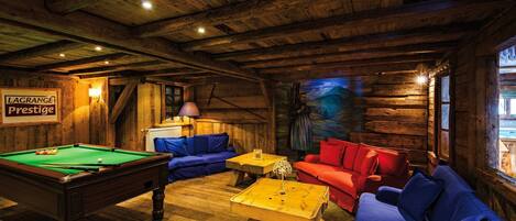 Interno di una baita in legno con tavolo da biliardo, divani blu e rossi e un quadro appeso al muro.