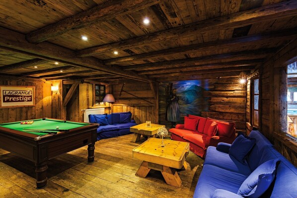 A wooden cabin interior with a pool table, blue and red sofas, and a painting on the wall.