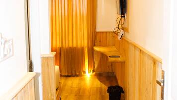 A narrow hallway with wooden flooring, a wooden bench, a black trash bin, and a window with curtains.