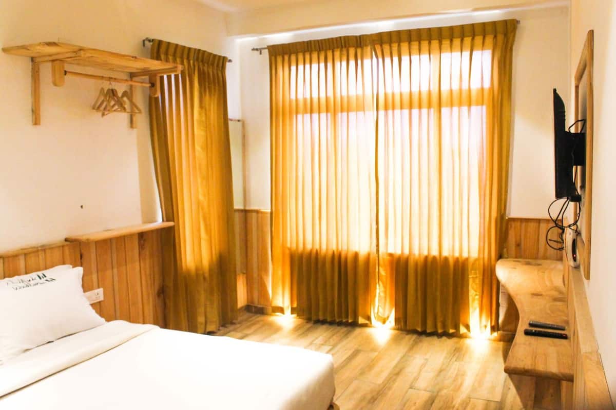 Une chambre d’hôtel avec un lit, une tête de lit en bois, une étagère en bois, un téléviseur fixé au mur et une fenêtre avec des rideaux.