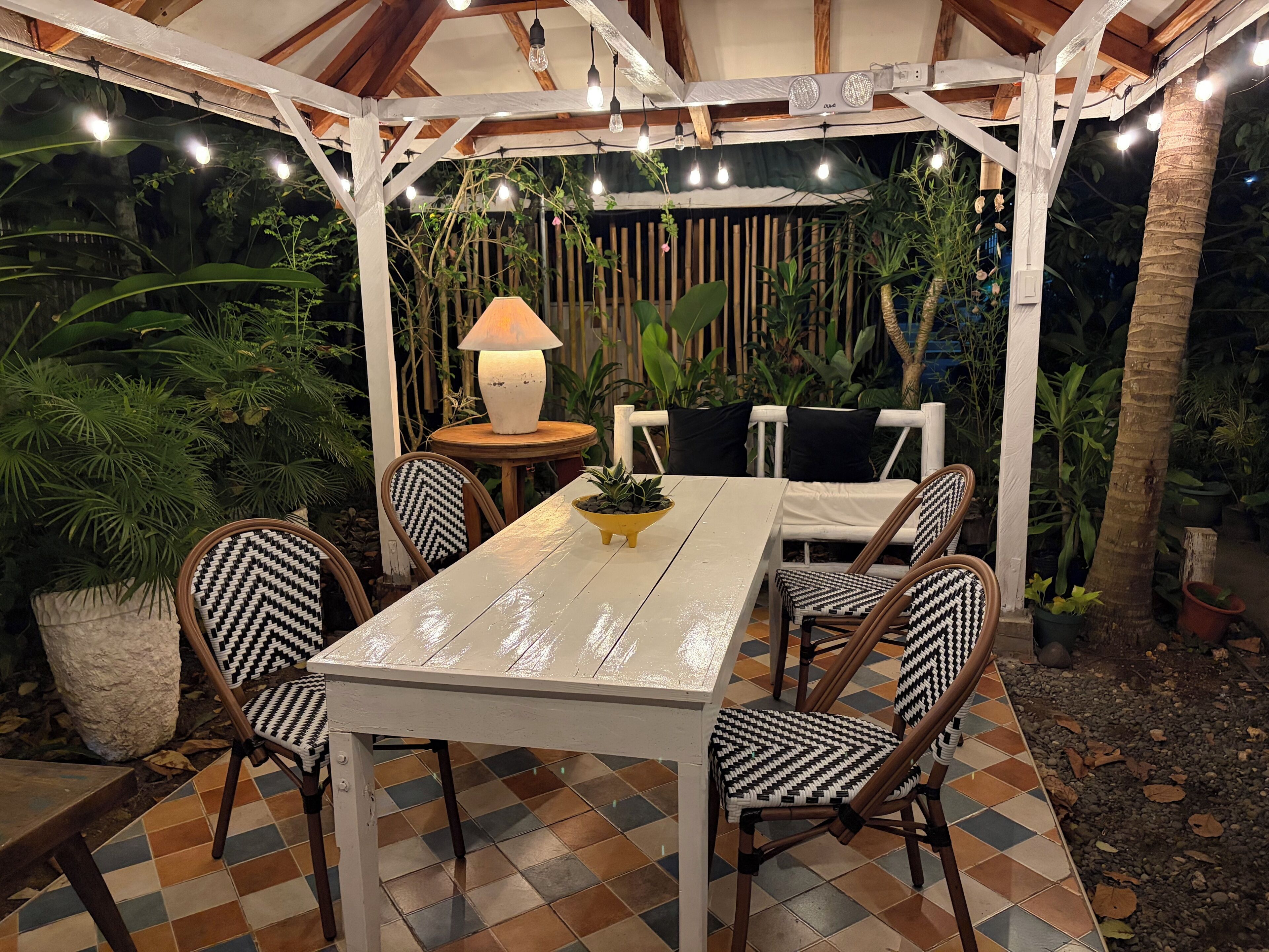 An outdoor patio with a white table and chairs, surrounded by plants and a wooden fence.