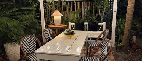 Un patio al aire libre con una mesa y sillas blancas, rodeado de plantas y una cerca de madera.