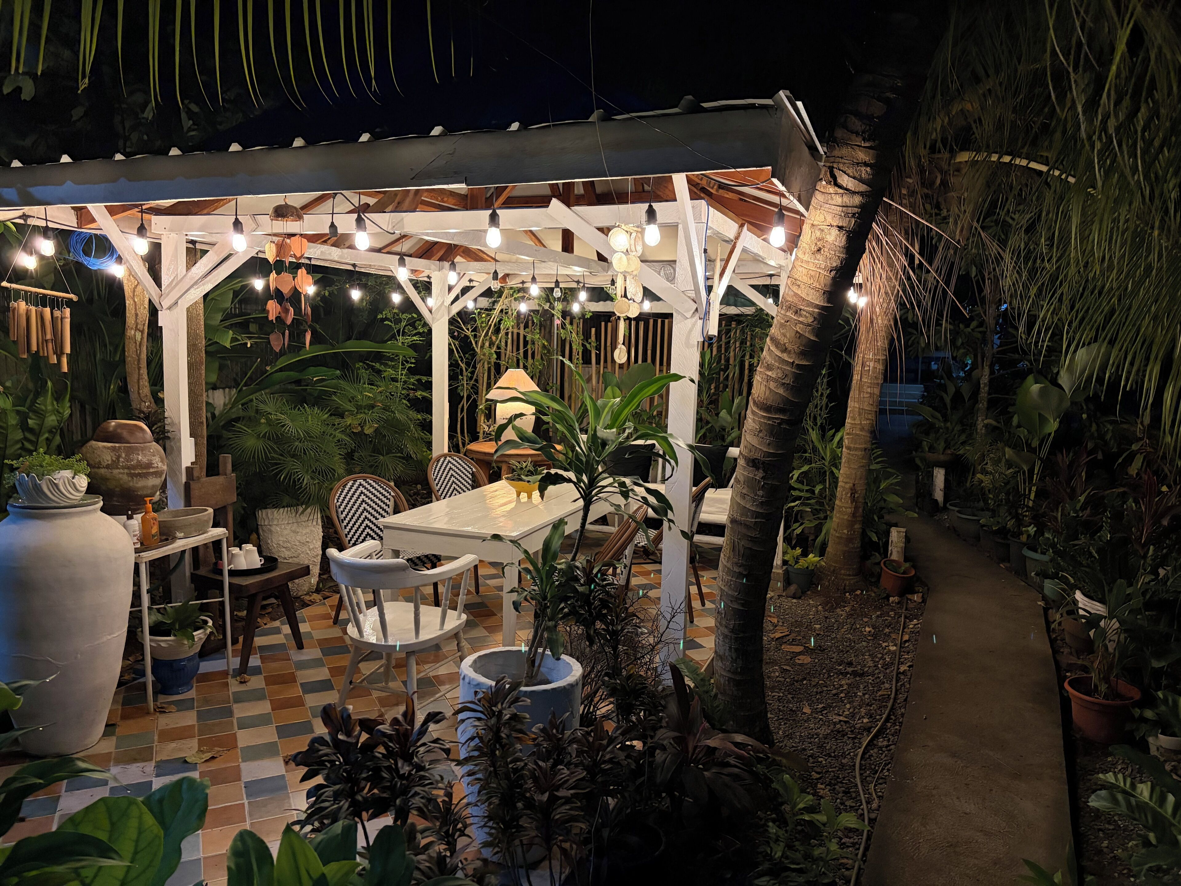 Eine Terrasse im Freien mit Schachbrettmuster-Boden, einem überdachten Sitzbereich mit Stühlen und Tisch sowie verschiedenen Blumentöpfen.