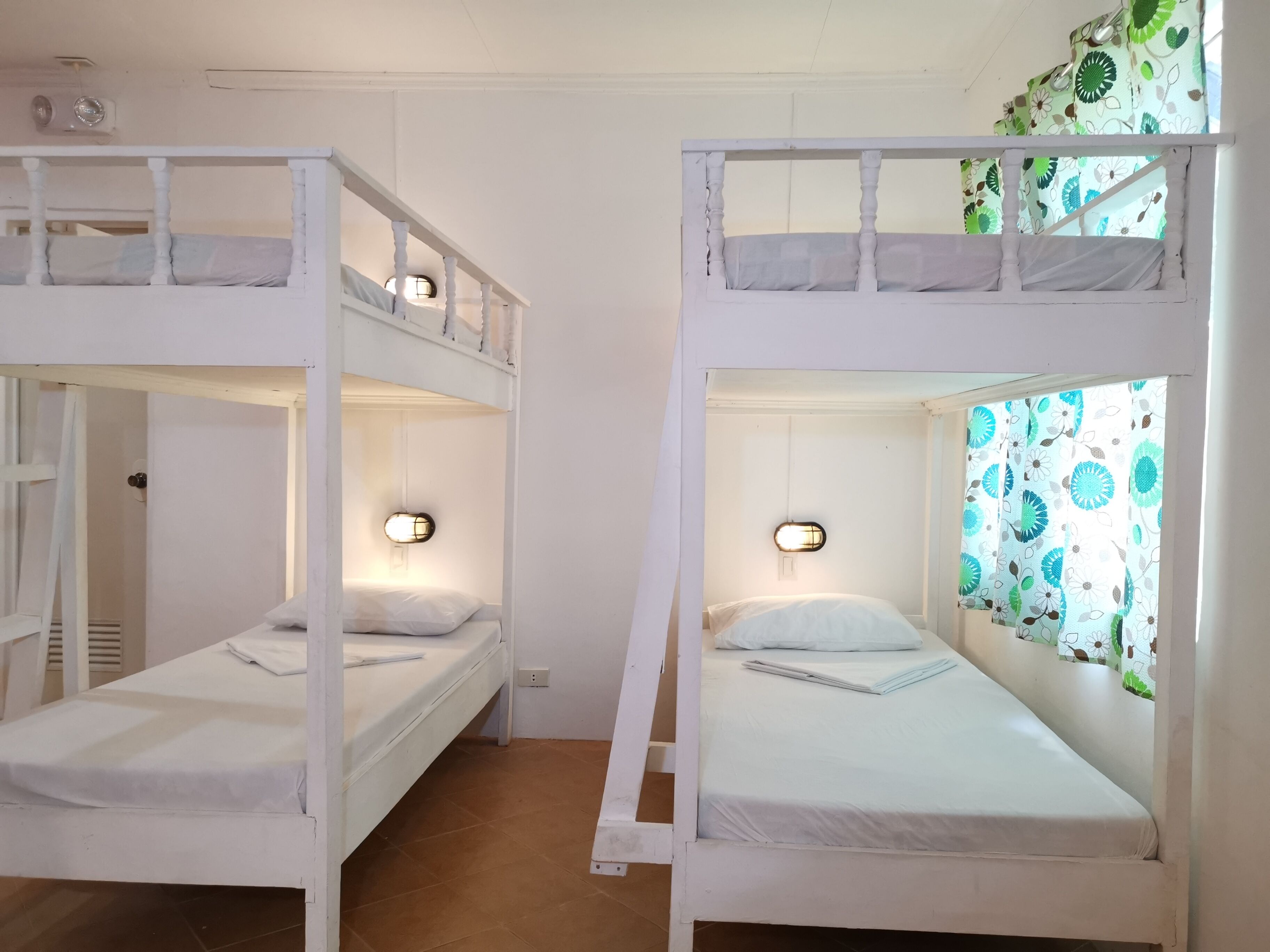Two bunk beds with white bedding, a wall-mounted light fixture, and a window with floral curtains.