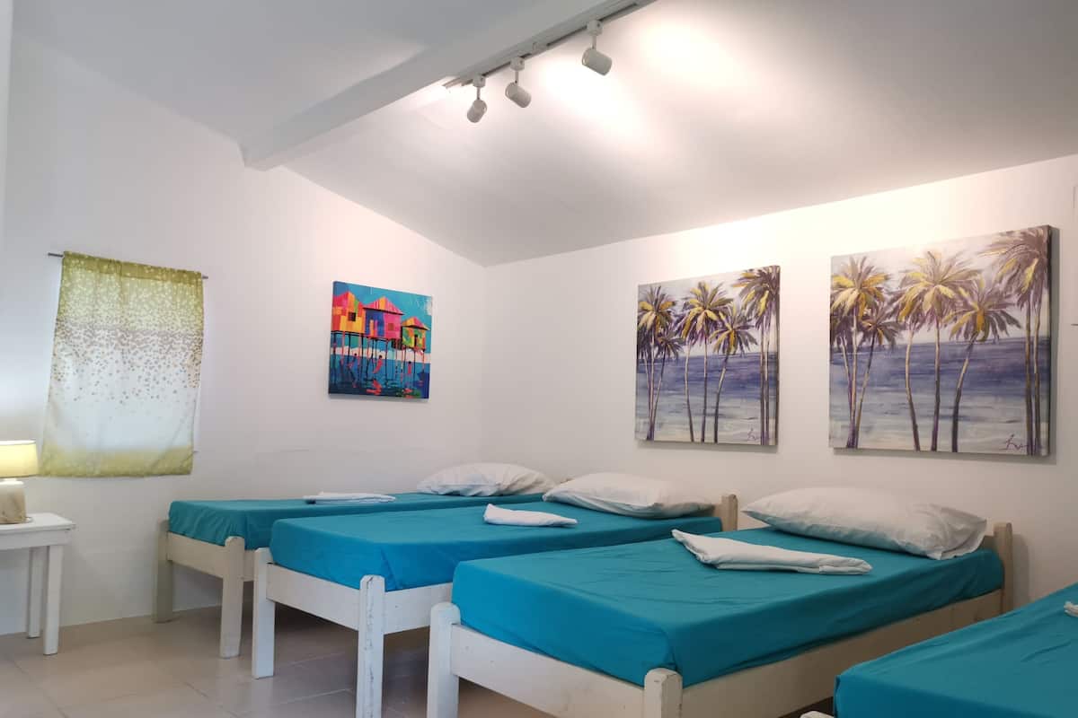 Una habitación con tres camas, cada una con colcha azul y almohadas blancas. Hay dos cuadros en la pared: uno muestra casas coloridas y el otro, palmeras.