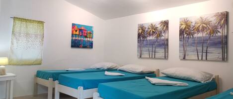A room with three beds, each with a blue bedspread and white pillows. There are two paintings on the wall, one depicting colorful houses and the other showing palm trees.