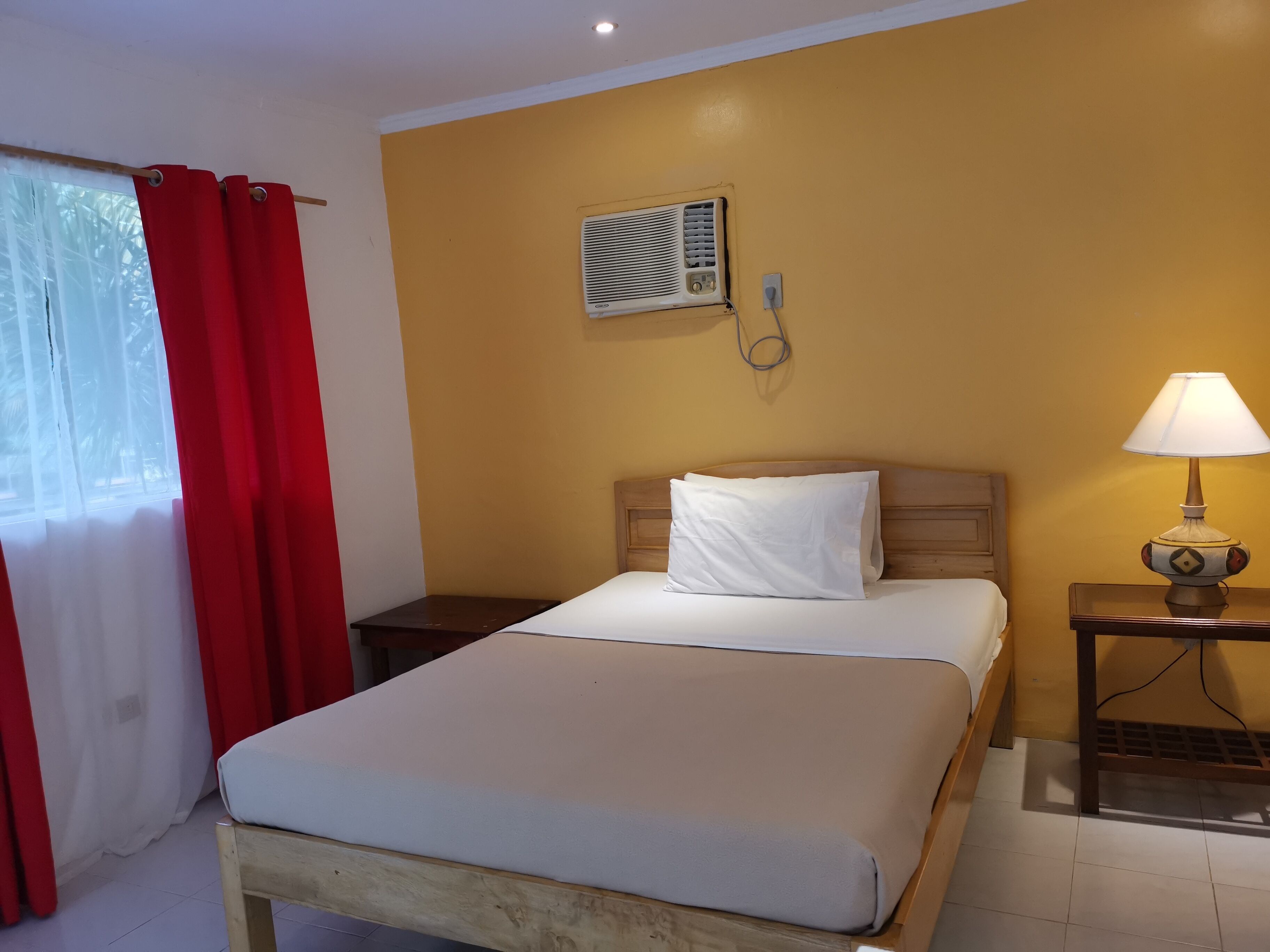 A hotel room with a bed, a bedside table with a lamp, an air conditioning unit, and a window with red curtains.