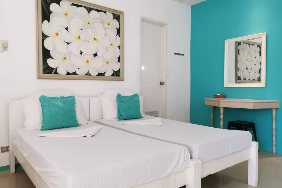 Una habitación con dos camas blancas, almohadones verde azulado, una puerta blanca, una pared de acento verde azulado, un lavabo blanco y una obra de arte floral enmarcada.