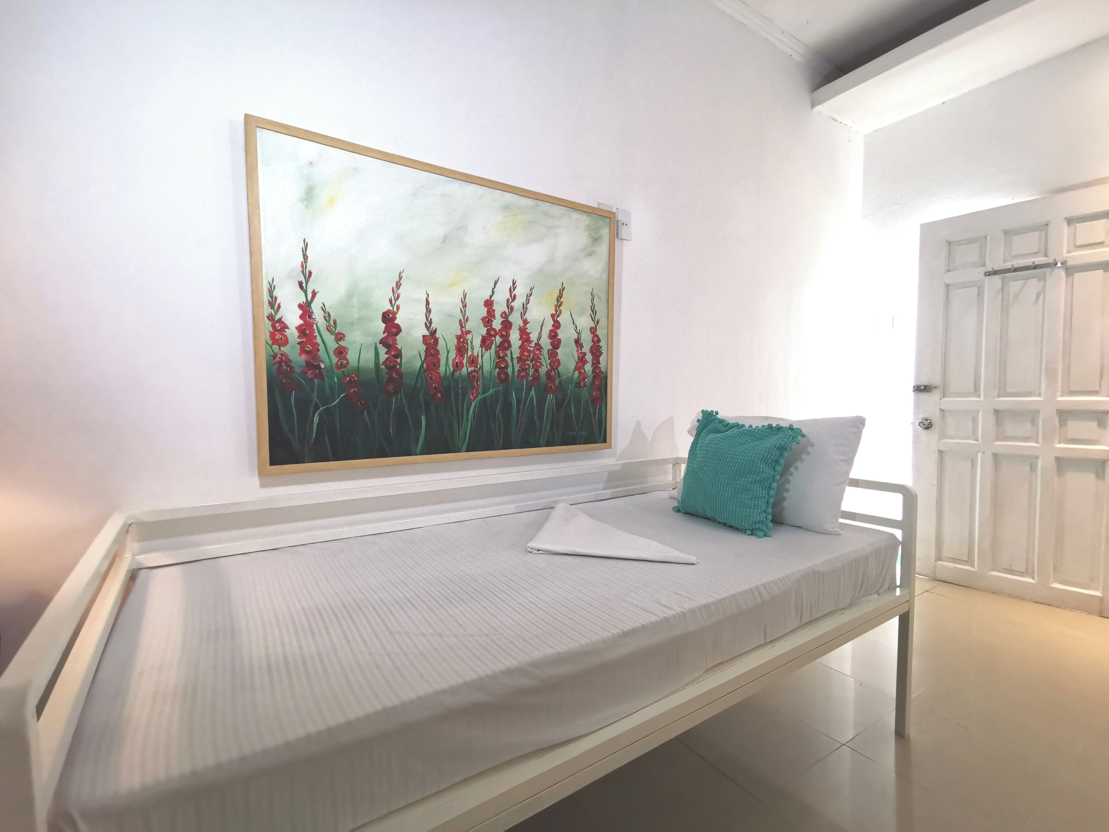 A white bed with a teal pillow, a white wall, and a framed painting of red flowers.