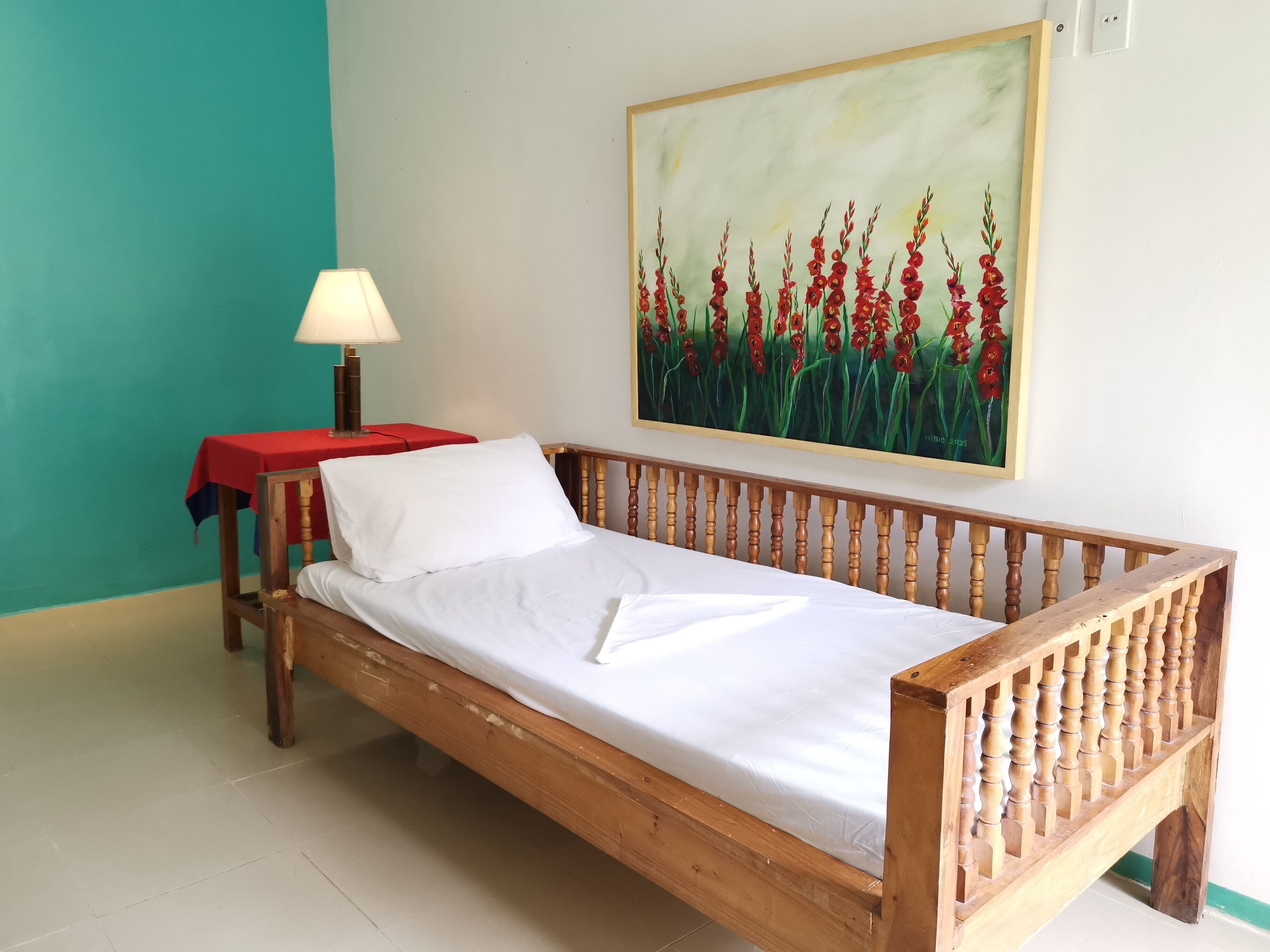 Una cama de madera con colchón y almohada blancos, una mesa roja con una lámpara, y un cuadro de flores rojas en la pared.