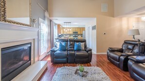 A living room with a fireplace, leather recliner, and a kitchen in the background. - Comfort and Style on the Colonnade (Rex)