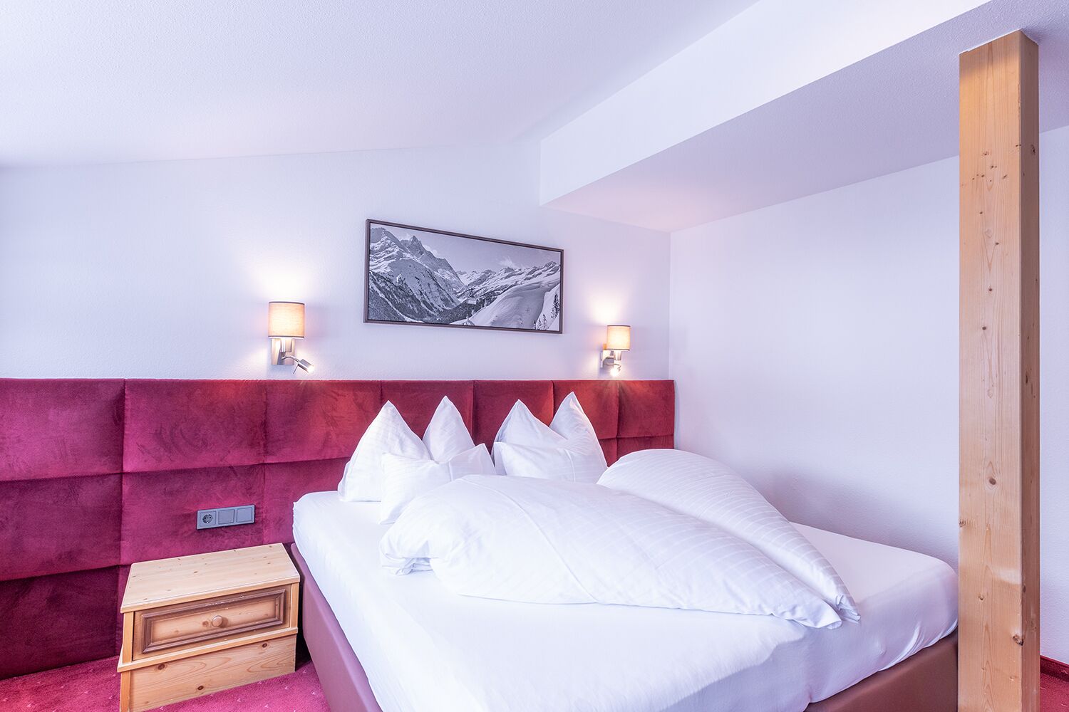 Un dormitorio con cabecera roja, ropa de cama blanca, dos lámparas de noche, una mesita de noche de madera y un cuadro en la pared.