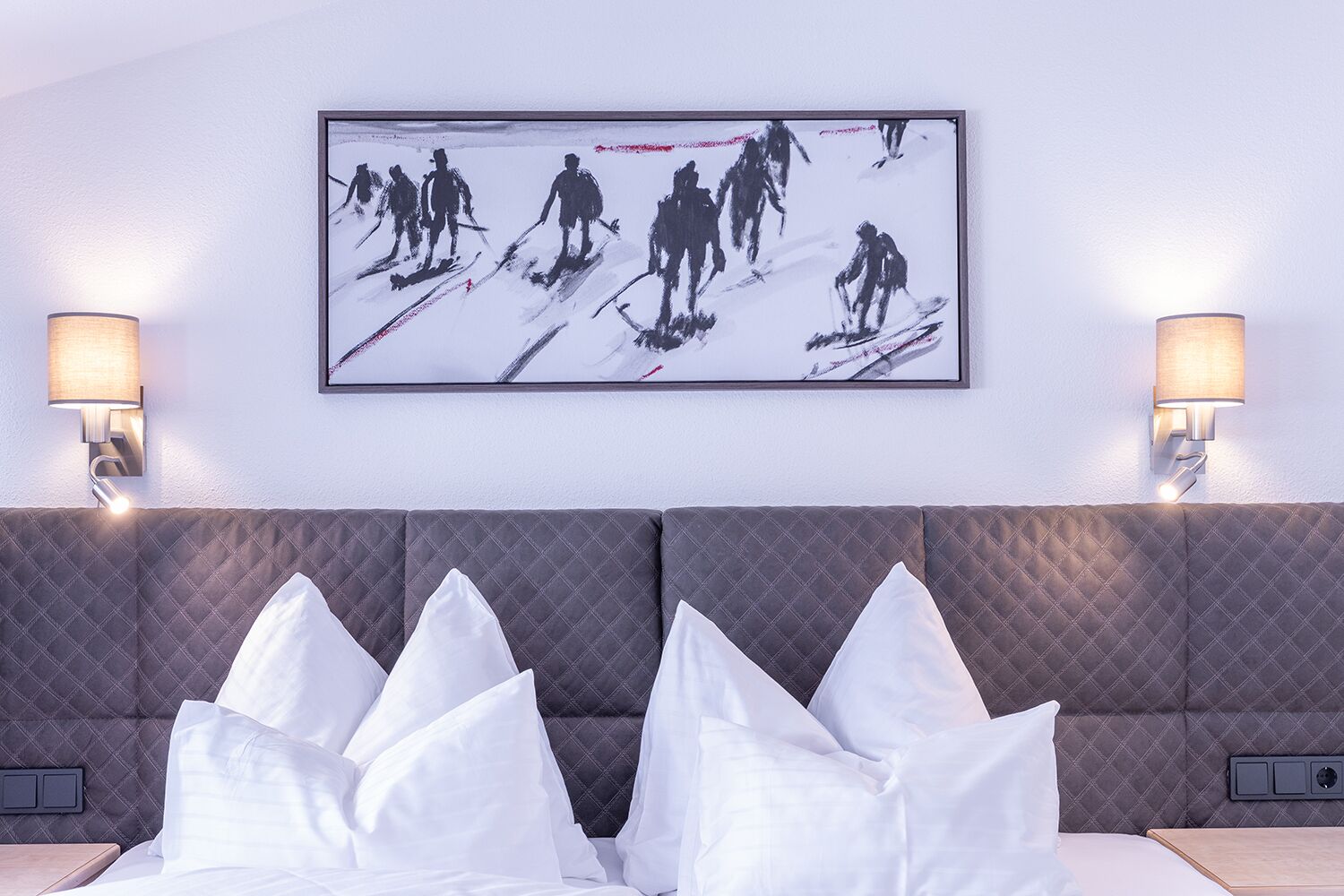 A hotel room with a large bed, a quilted headboard, two wall-mounted lamps, and a framed picture of skiers on the wall.