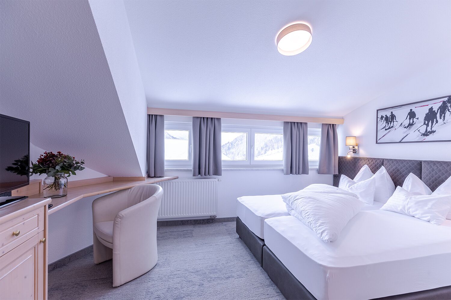 A modern hotel room with a bed, a desk, a chair, a TV, and a window with curtains.