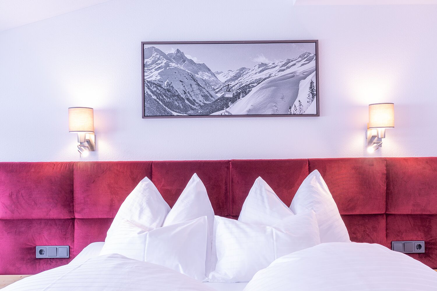 A hotel room with a red upholstered headboard, white bedding, and a framed mountain landscape picture on the wall.