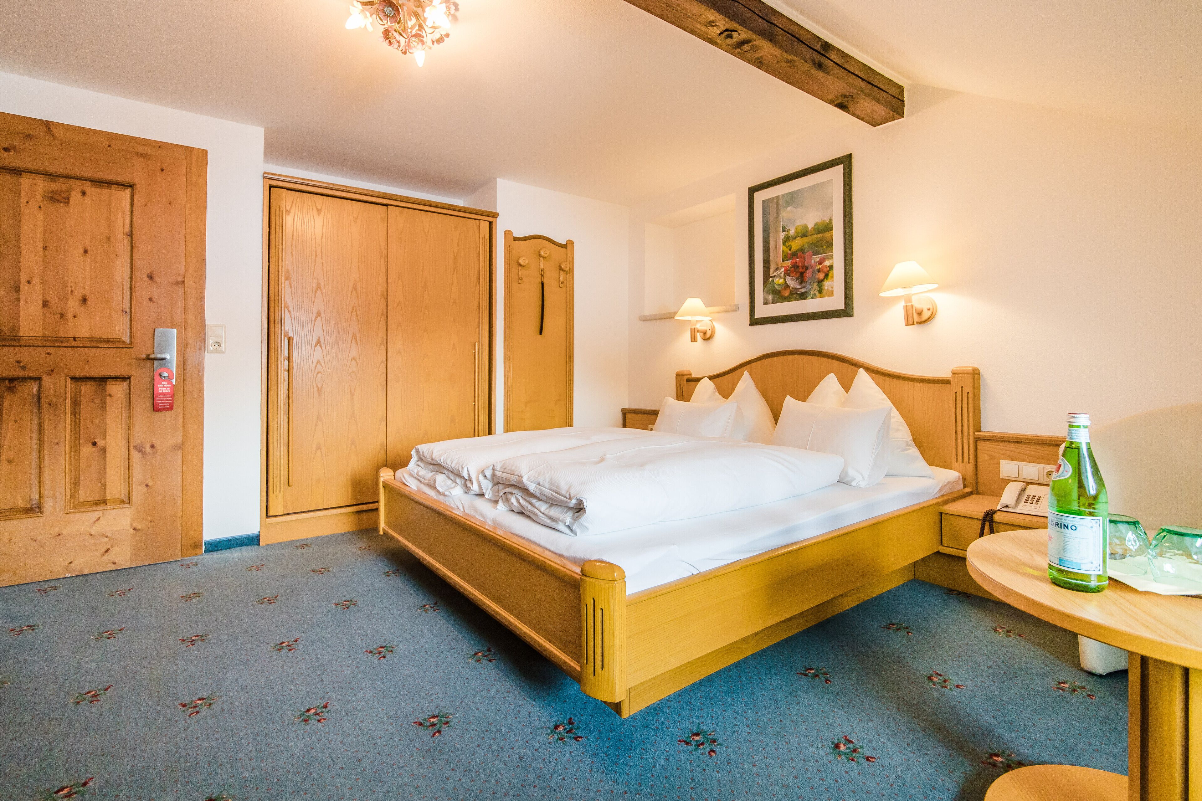 A hotel room with a wooden bed, a small bedside table, a lamp, and a painting on the wall.