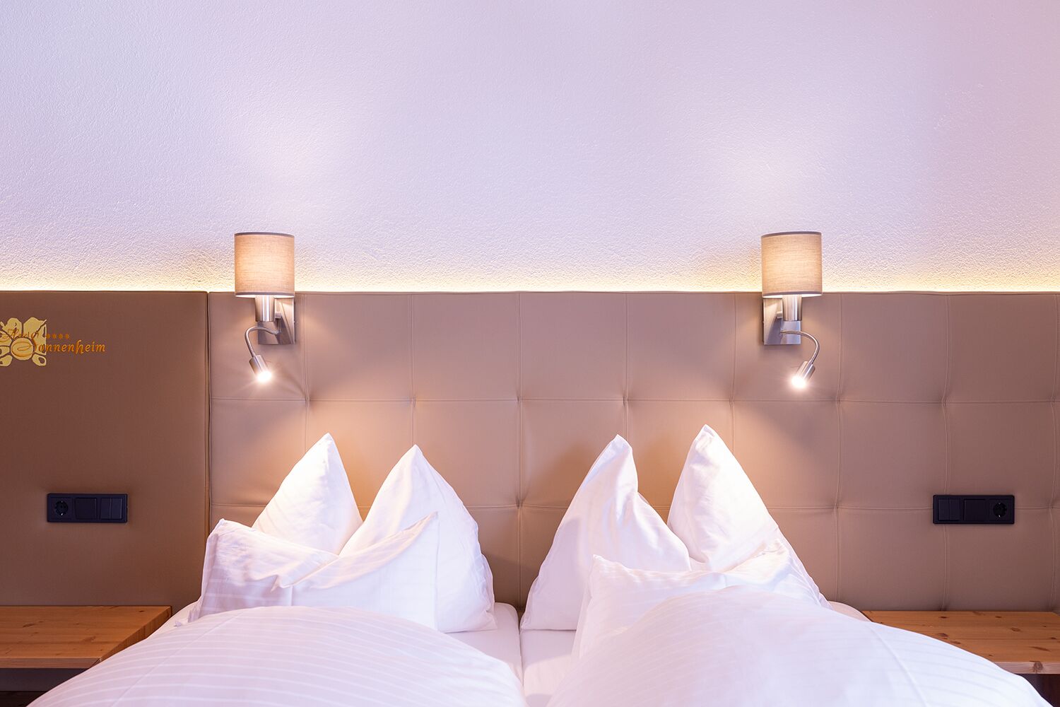 Habitación de hotel con dos camas, cabecero acolchado, lámparas fijadas a la pared y mesitas de noche de madera.