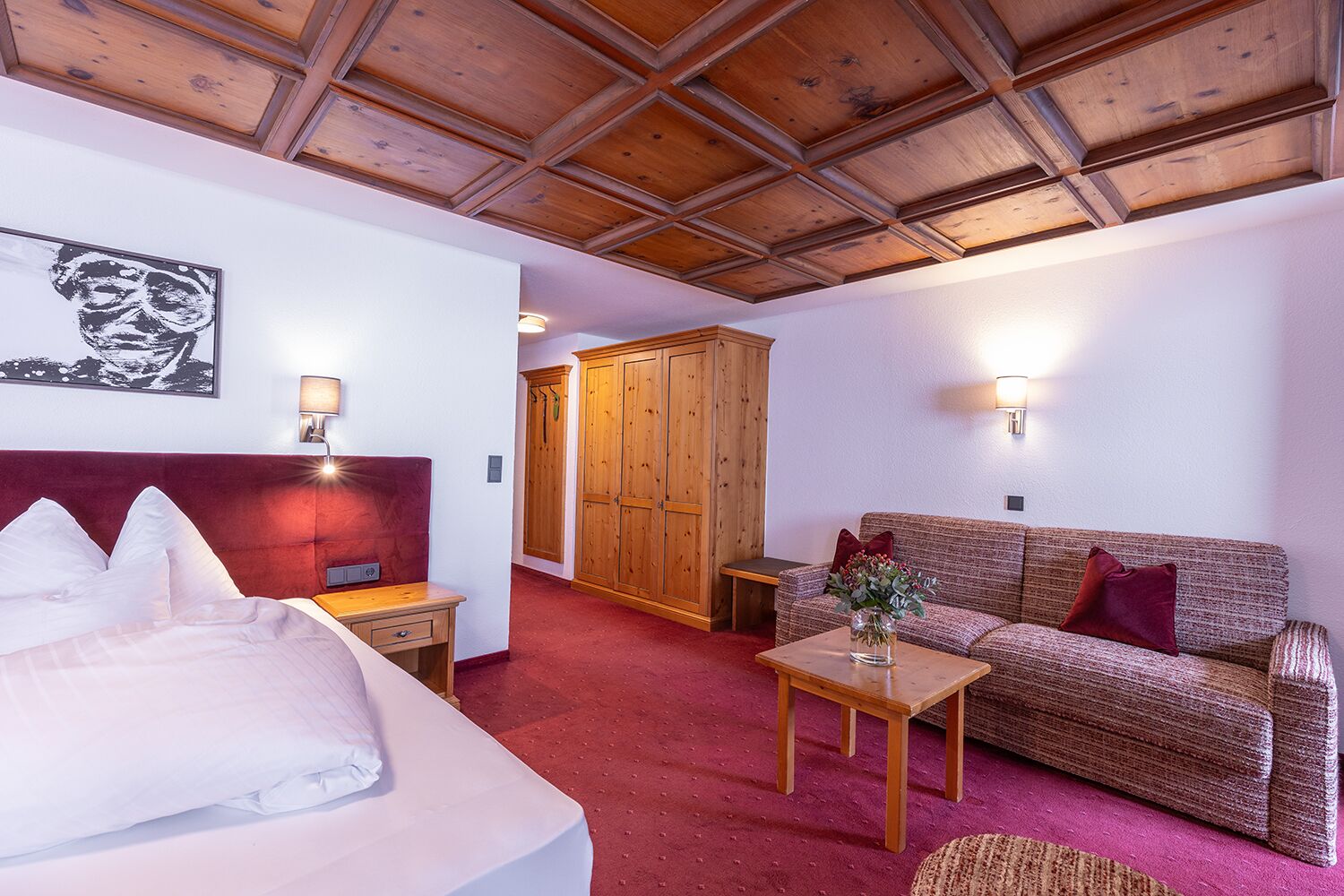 A hotel room with a bed, a wooden headboard, a sofa, a small table with a vase, and a wardrobe.