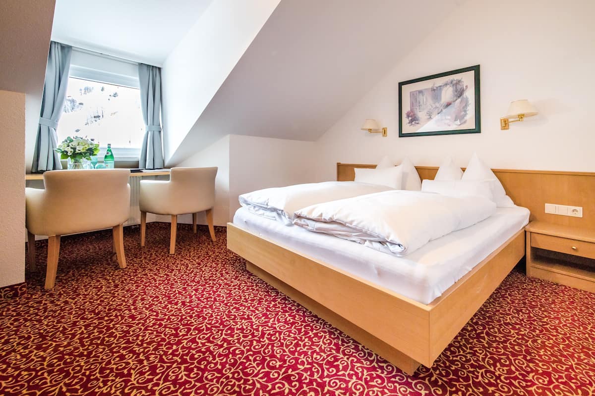 A hotel room with a bed, two bedside tables, a desk with chairs, a window with curtains, and a patterned carpet.