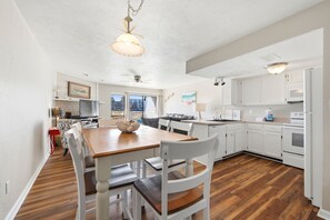 A kitchen with white cabinets, a dining table with chairs, a ceiling fan, and a view of the outside through a window.