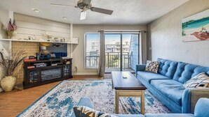 A spacious living room with a blue sofa, a coffee table, and a ceiling fan. There is a large window with a view of the city.