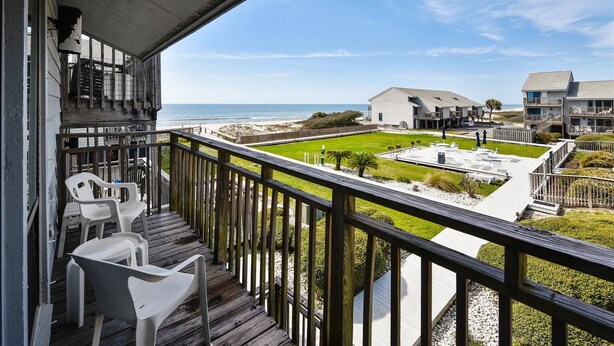 A balcony with a view of the beach and ocean, wooden deck, and outdoor seating area.