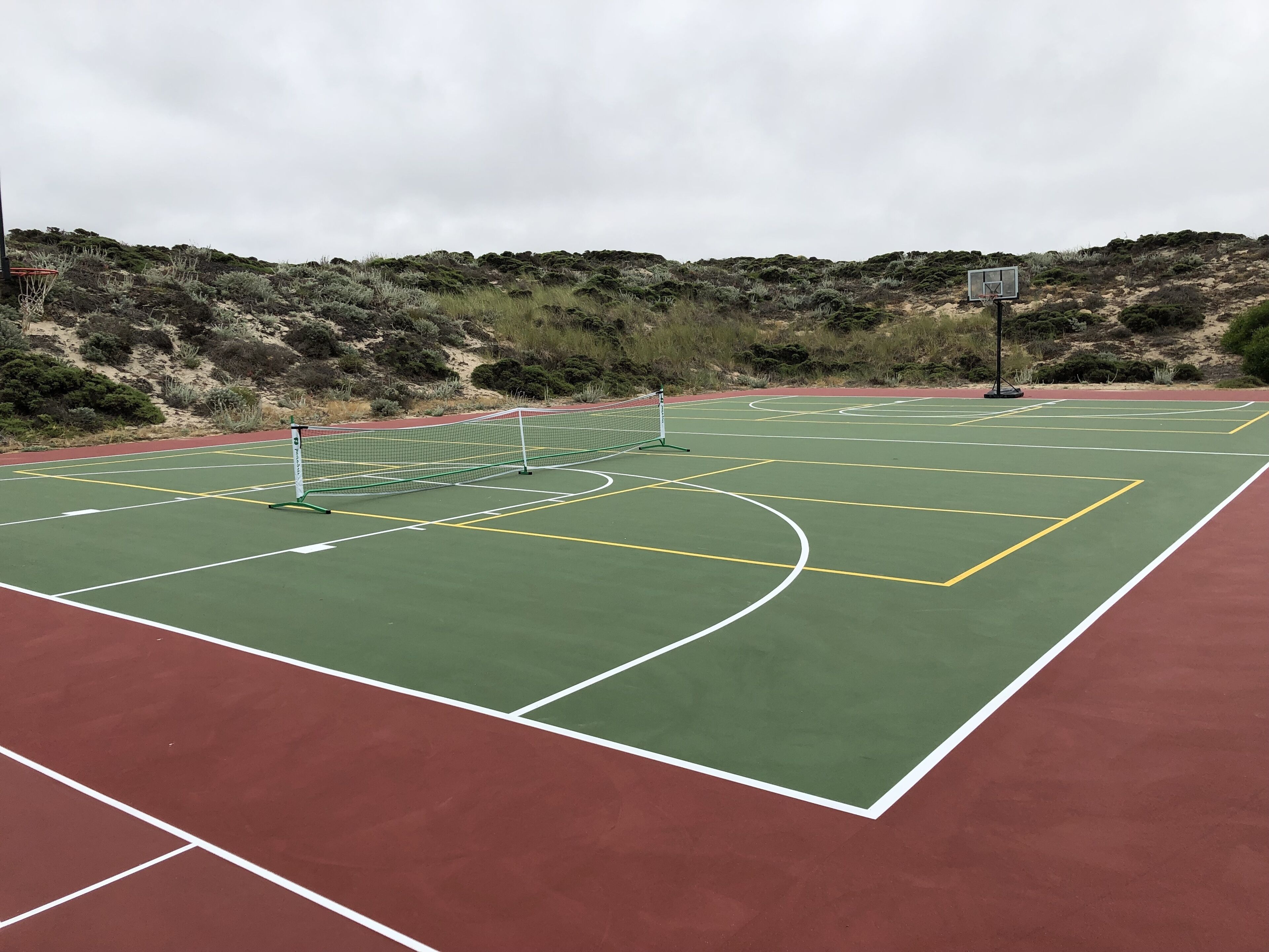 Een basketbalveld buiten met een net, omgeven door groene en rode vloerbedekking, en een heuvelachtig landschap op de achtergrond.
